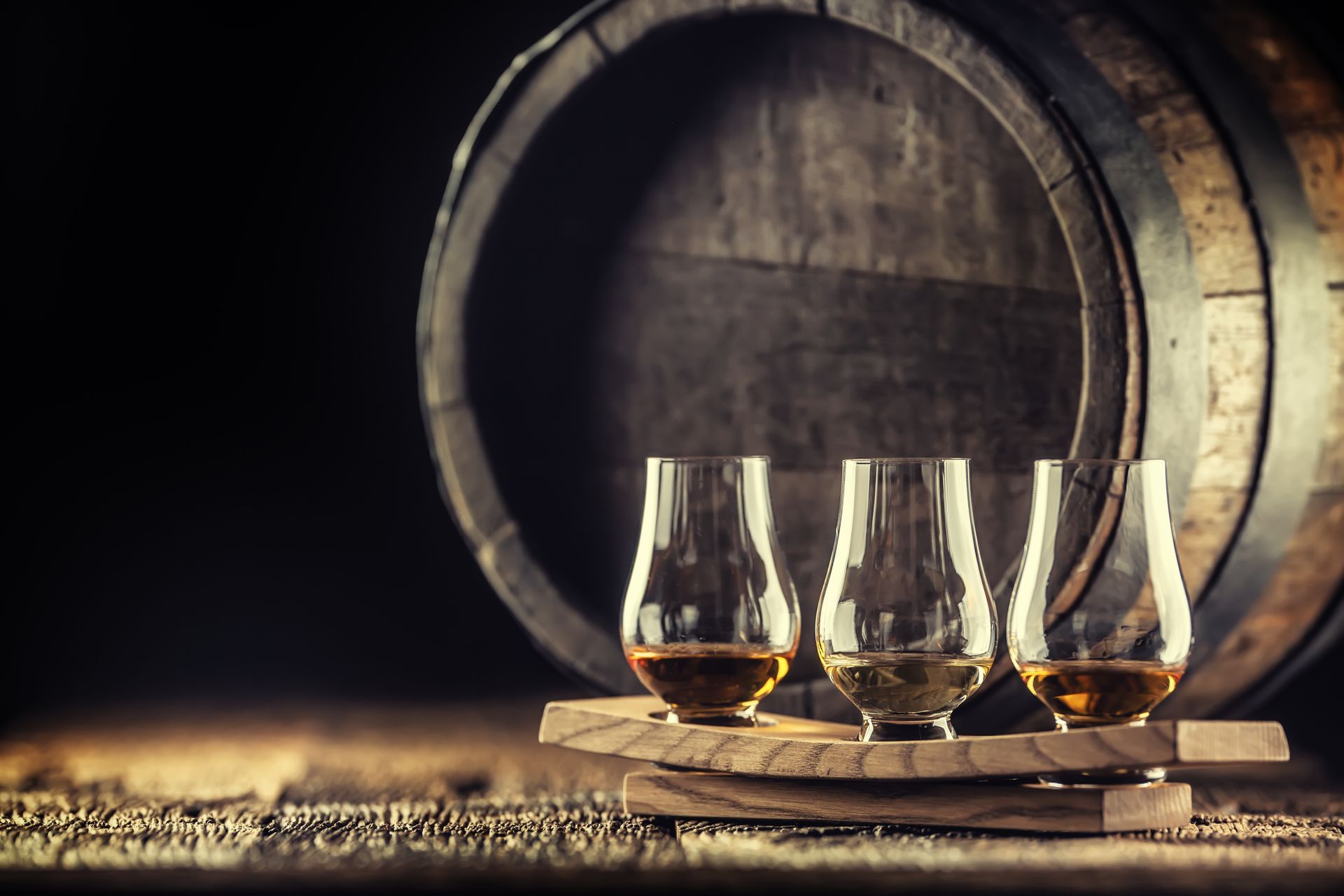 Three glasses of whiskey are sitting on a wooden tray in front of a wooden barrel.