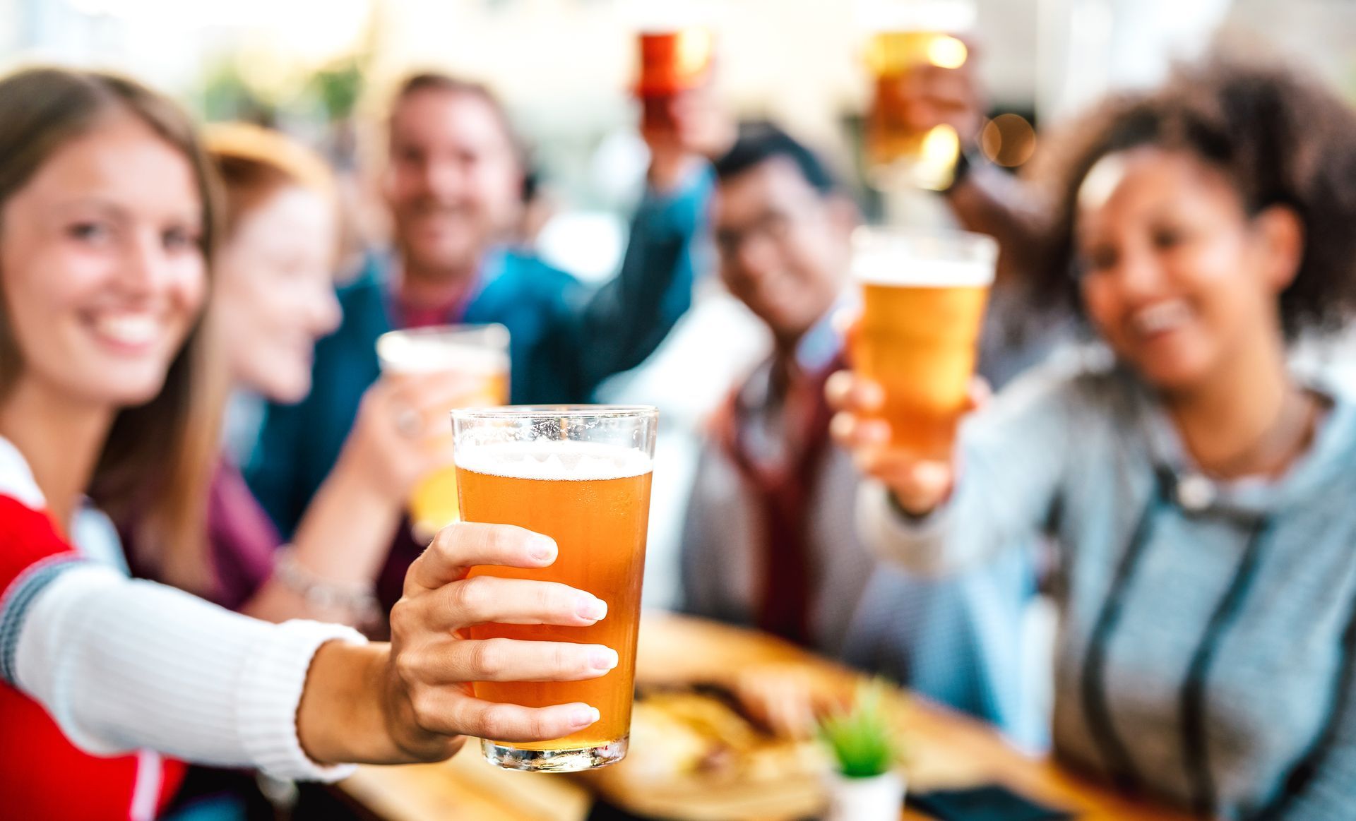 Group of friends at a table, toasting with beer glasses. Smiling faces, outdoor setting.