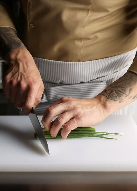 A man with a tattoo on his arm is cutting green onions on a cutting board.