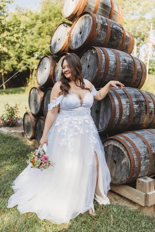 A plus size bride in a wedding dress is standing in front of a pile of wine barrels.