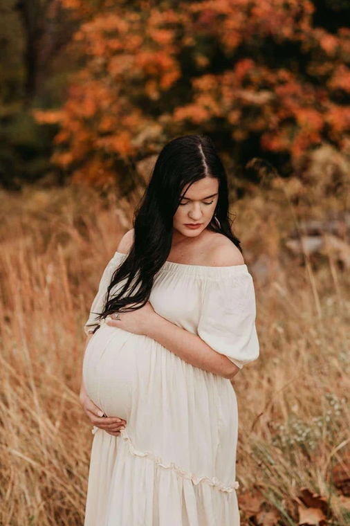 A pregnant woman in a white dress is standing in a field holding her belly.