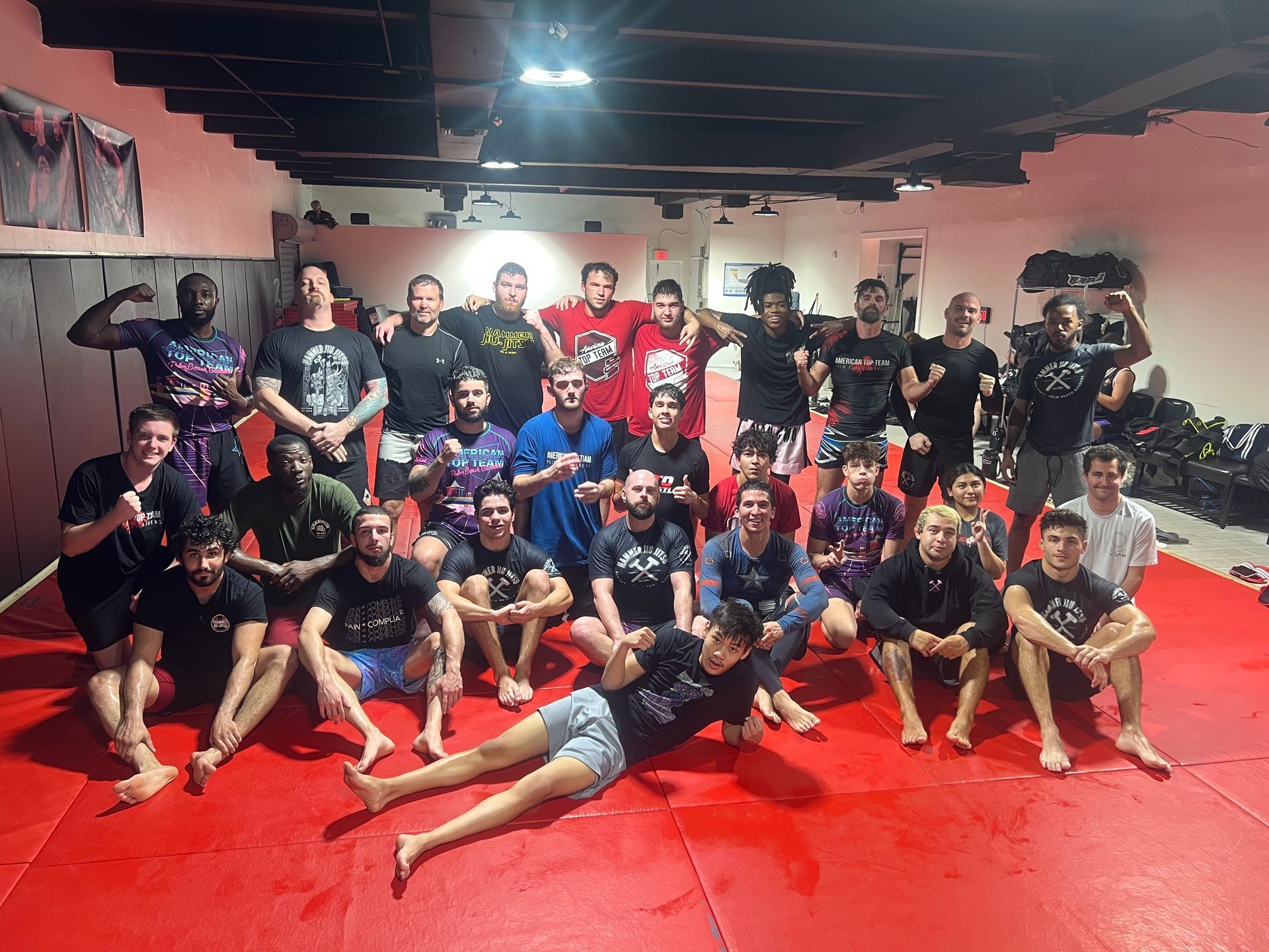 Group of people in gym, posing on red mats. Many are shirtless, wearing athletic wear.