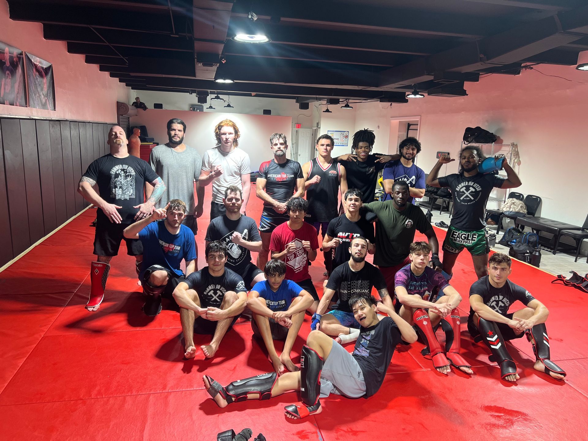 A group of people posing together in a fitness studio, most making hand gestures, with others training in the background.
