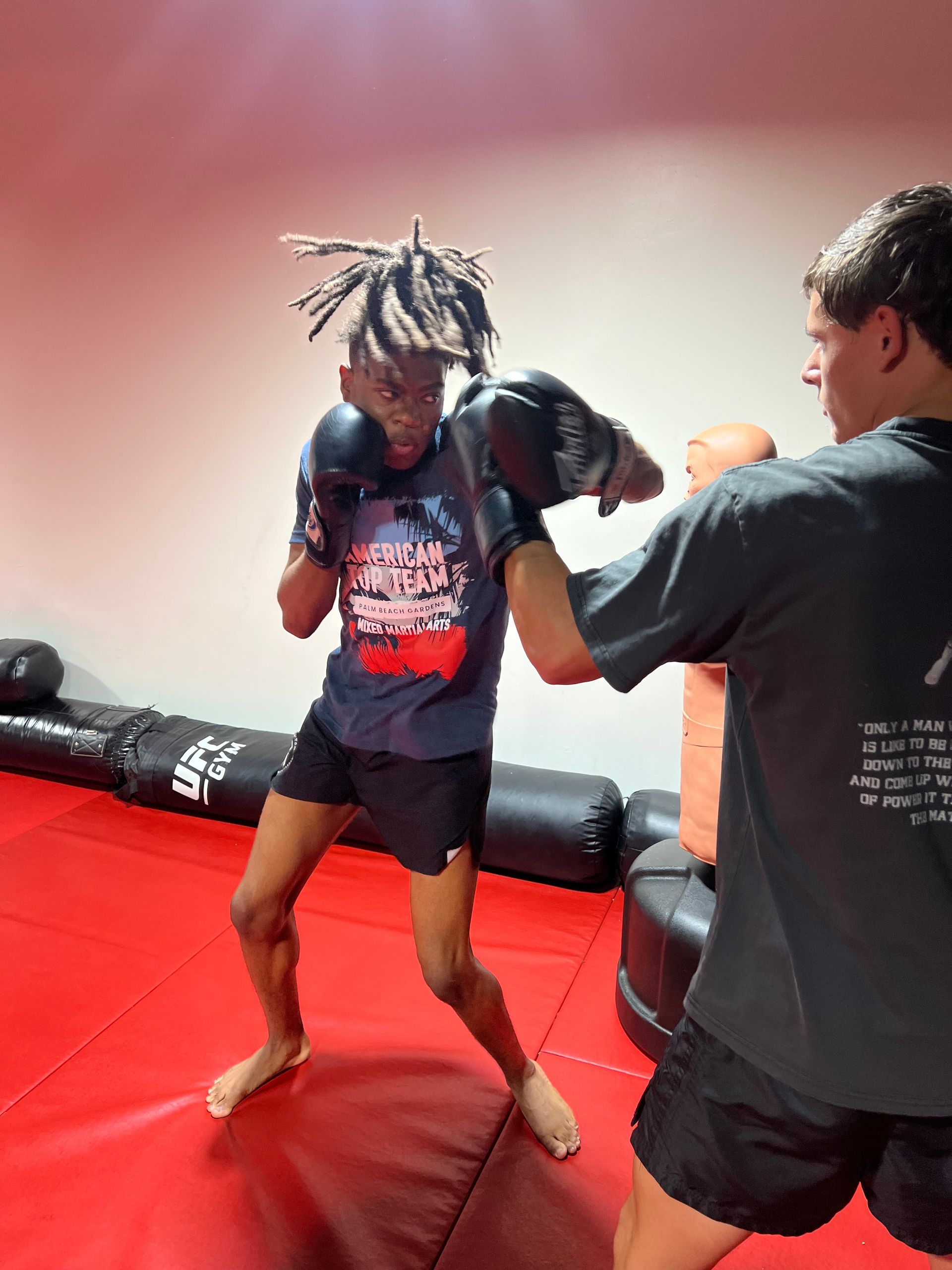 Person in boxing stance sparring with trainer in a gym, wearing gloves and shorts.