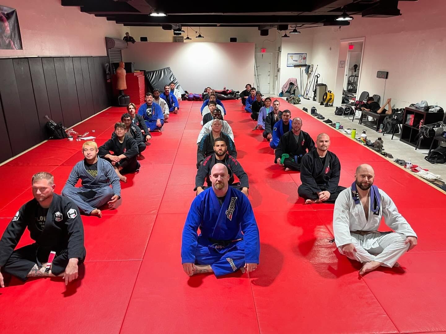 A group of people wearing martial arts uniforms sitting on a large red mat in a training studio.