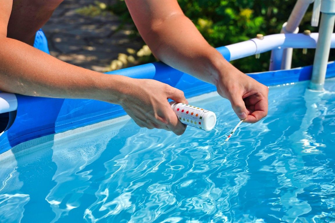 Hands cleaning a blue pool with a brush in bright sunlight