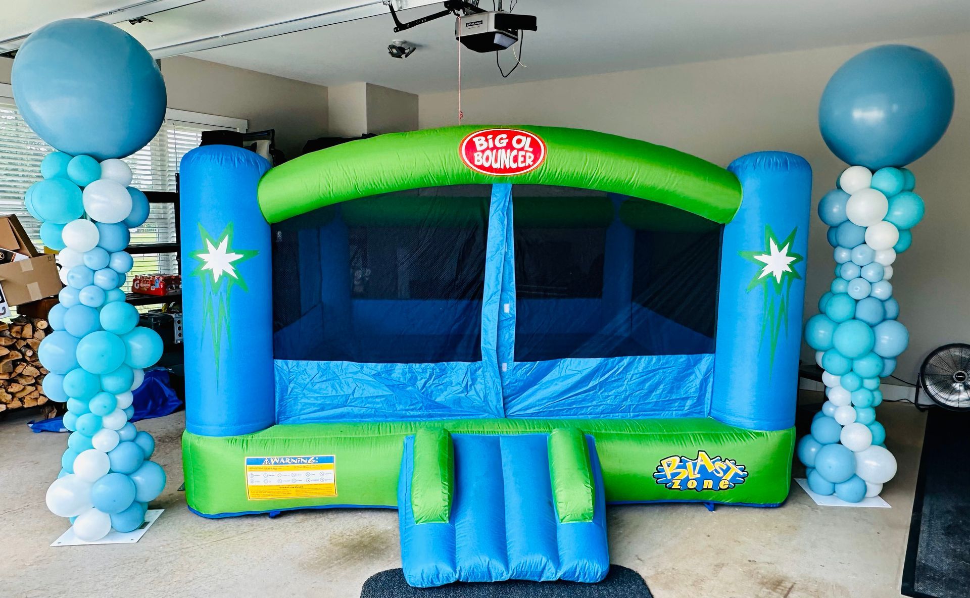 A blue and green bouncy house is surrounded by blue and white balloons.