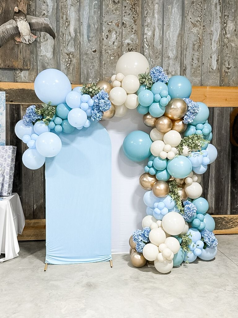 A blue , white and gold balloon arch is sitting in front of a wooden wall.