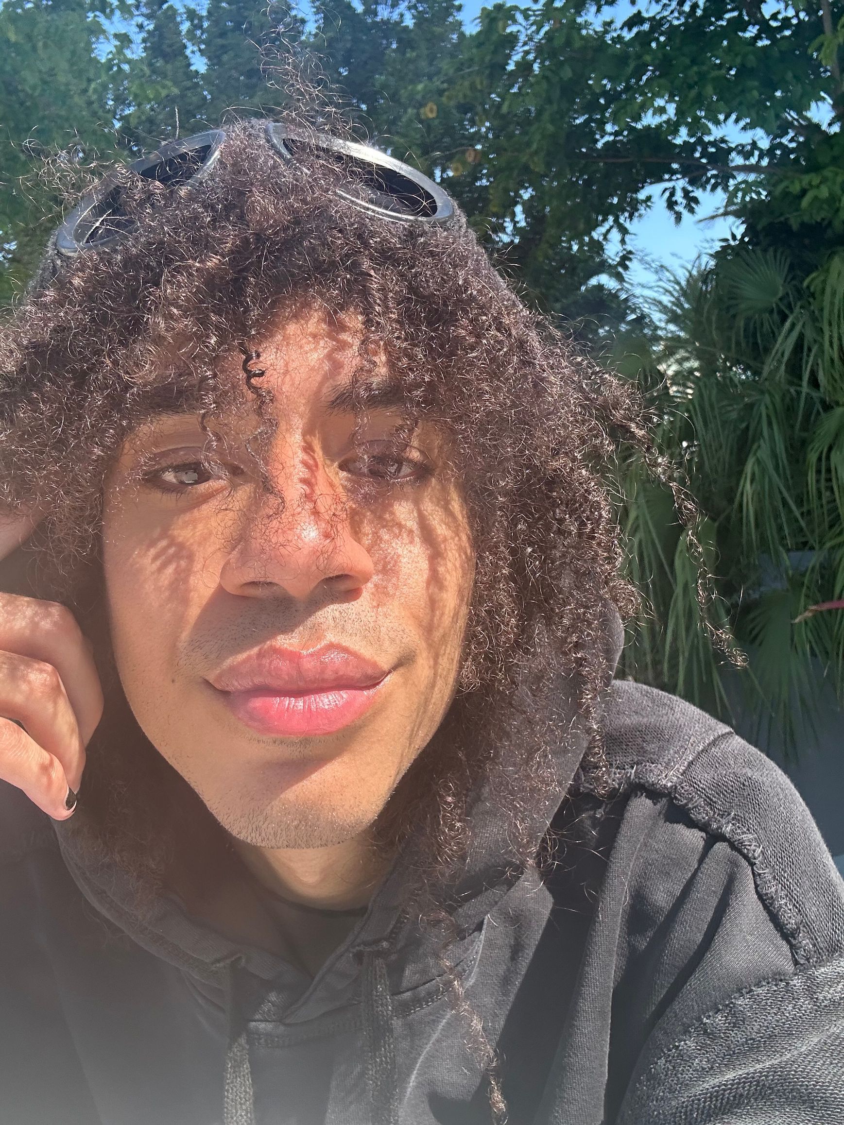 Man with curly hair, outdoors, in a hooded sweatshirt. Sunlit face, looking toward the camera.