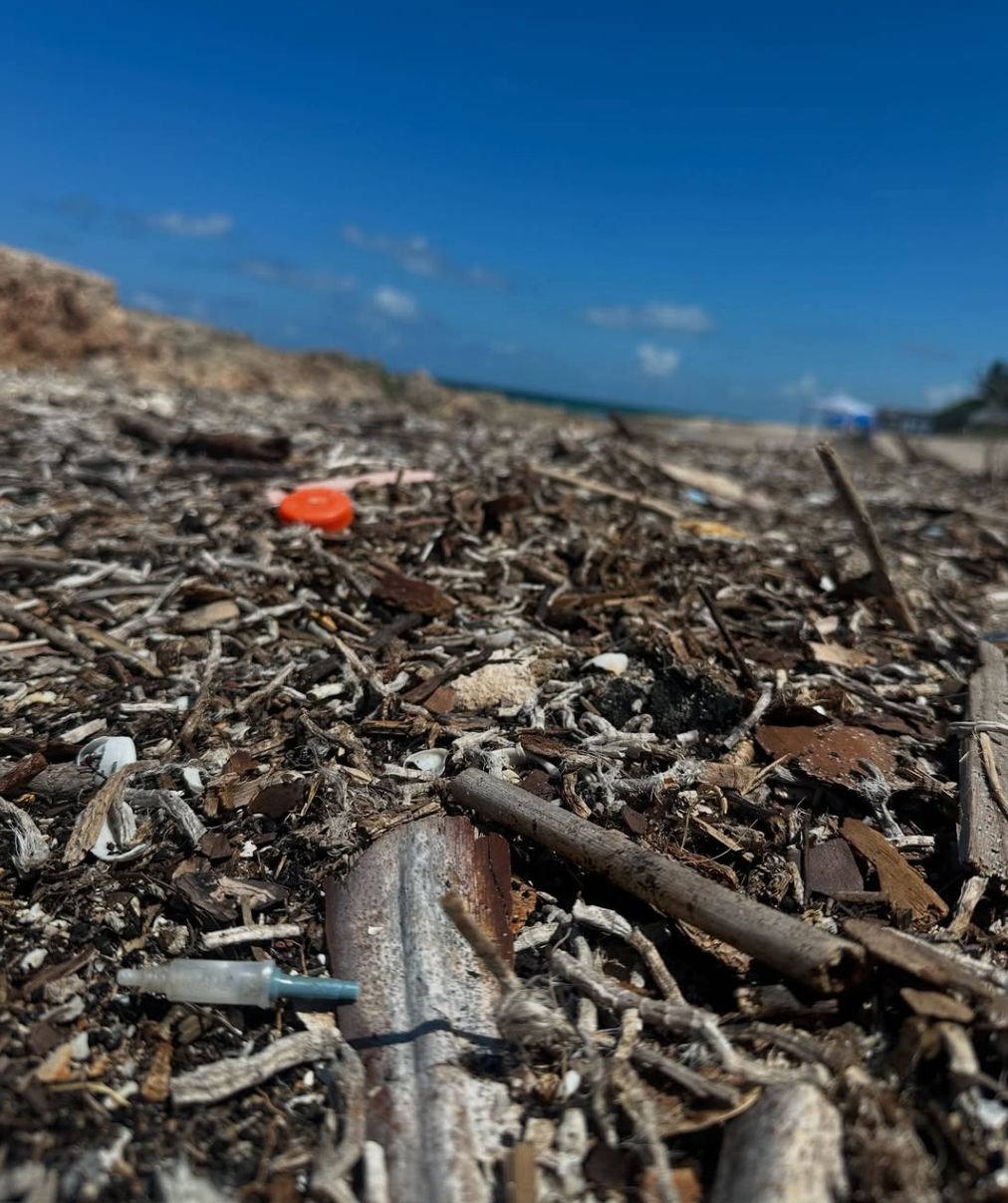 Close-up view of debris-strewn beach with a blue sky and ocean in the background. An orange bottle cap is visible amidst the debris.