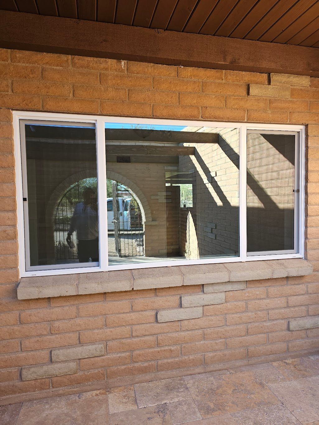 A white-framed window with a screen. The brick wall reflects a person standing outdoors with shadows.