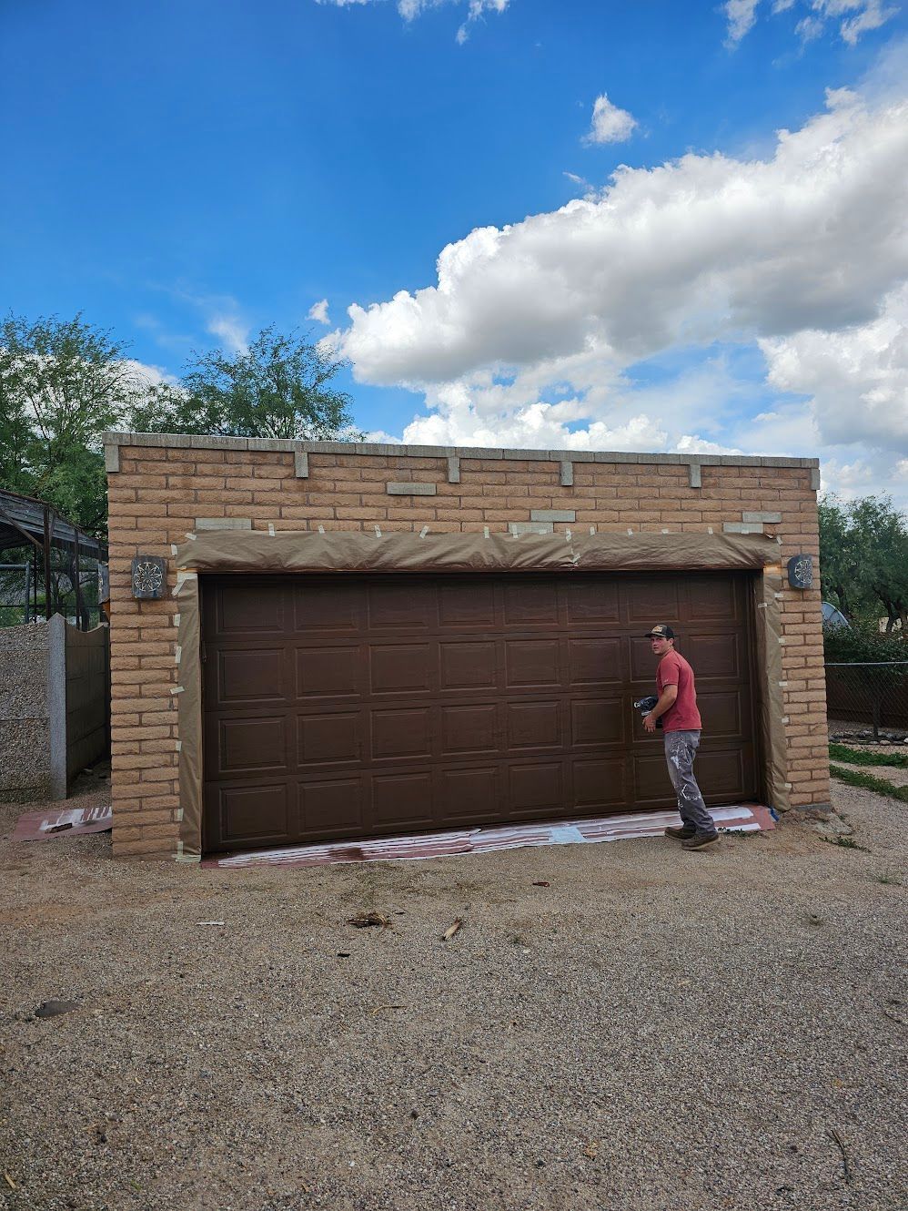 Man painting brown garage door on brick building under blue sky with clouds.