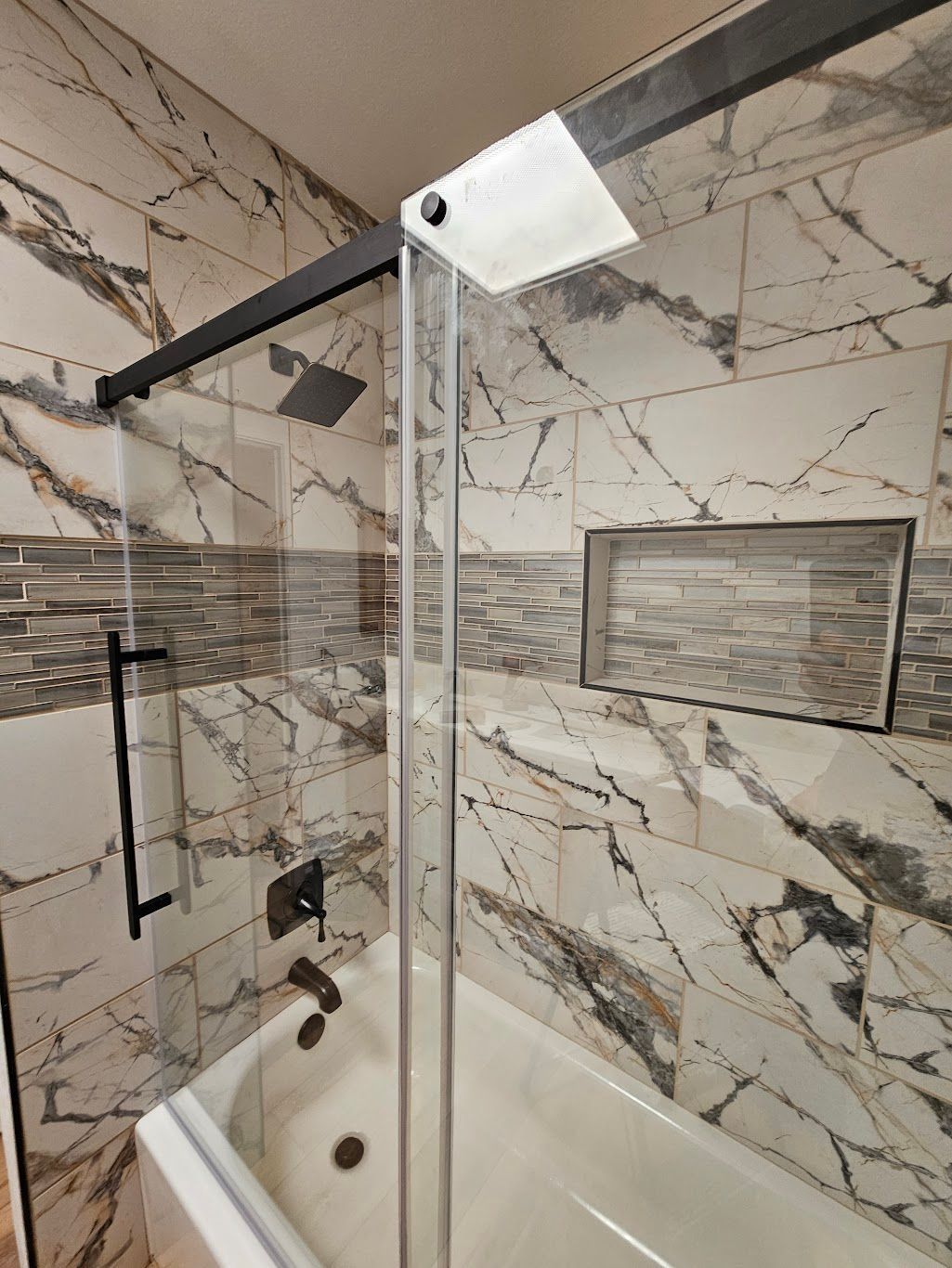 Shower with glass door, black hardware, white and marble-patterned tiles.