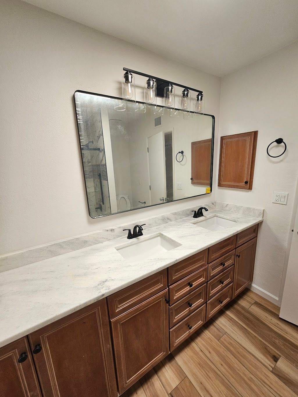 Bathroom vanity with marble countertop, brown cabinets, large mirror, and black fixtures.