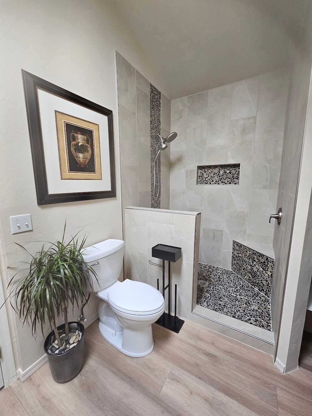 Bathroom with toilet, shower, artwork, and potted plant. Beige, gray, and brown tones.