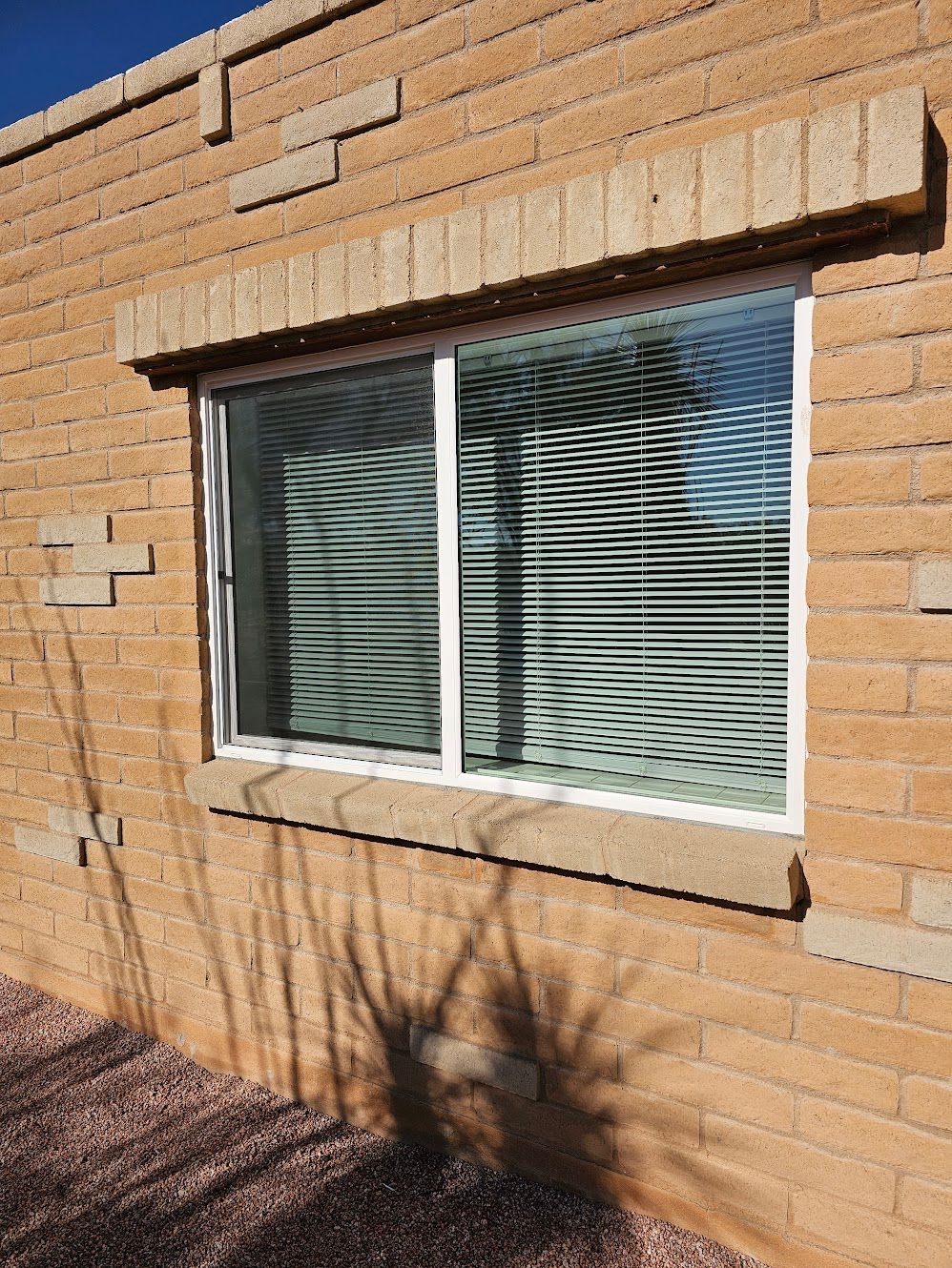 Window with white frame, horizontal blinds, on a tan brick wall with shadow.