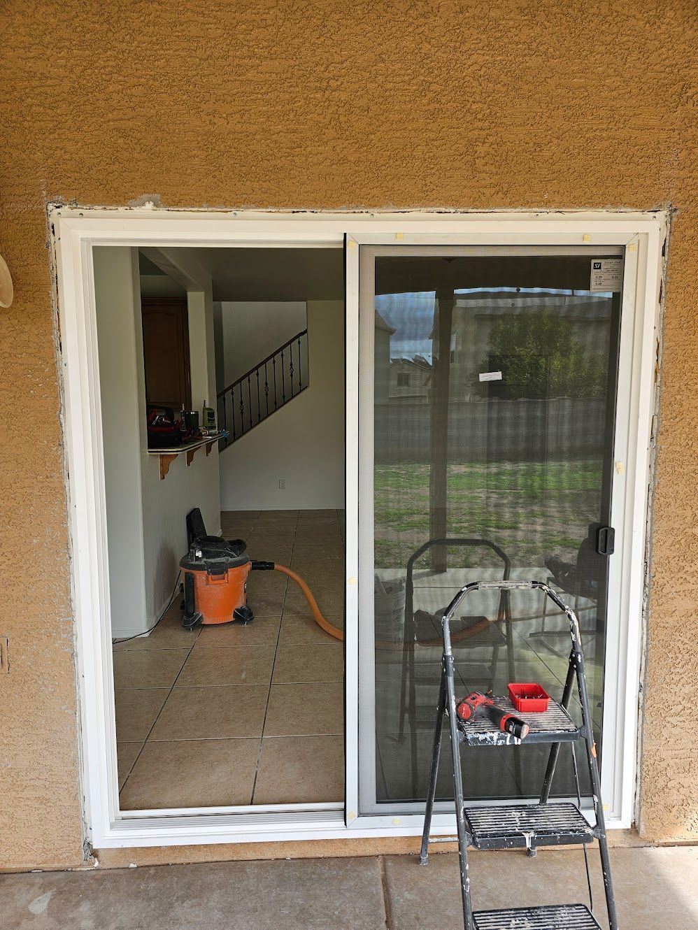 Sliding glass door frame being worked on, exterior view. White trim, open door, ladder, vacuum.