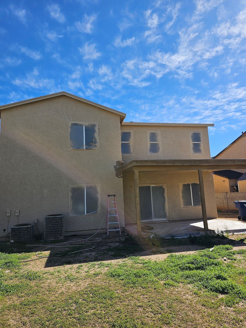 Back of a two-story beige stucco house with a covered patio. Windows are taped, ladder leans on the wall. Sunny, blue sky.