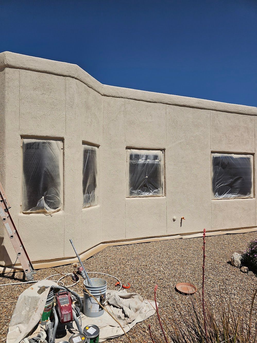Exterior of a stucco house with windows covered, paint supplies on the ground, and clear blue sky.