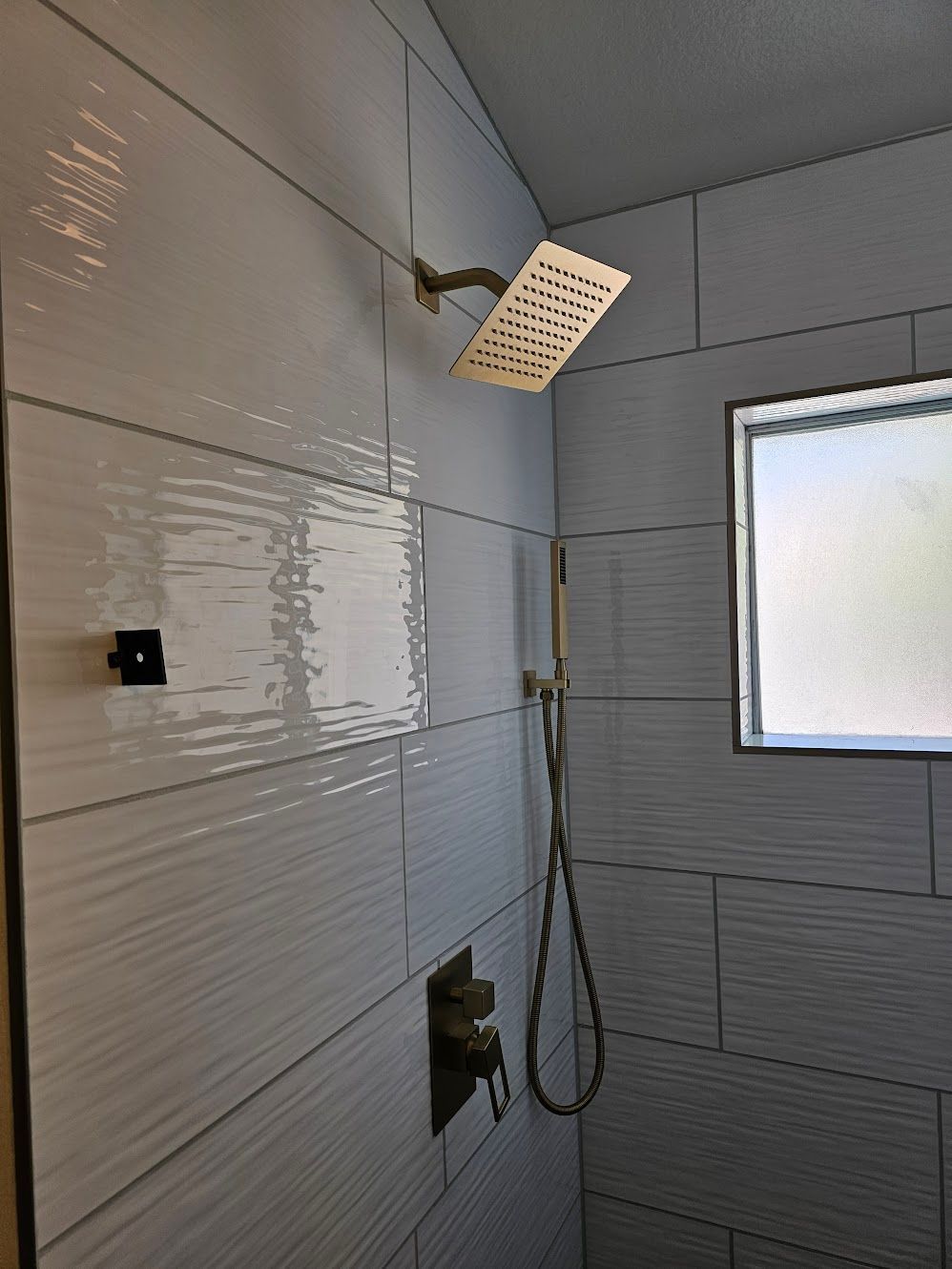 Shower with gold fixtures and textured white tiles. Small window on the right.