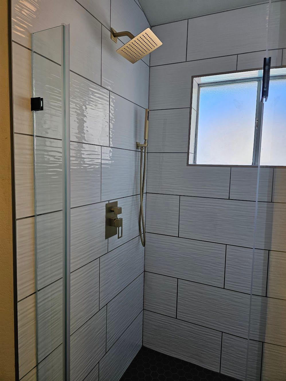 Shower with white textured tile walls, glass door, window, and gold shower fixtures.