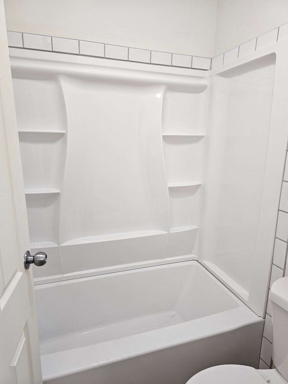 White bathtub and shower surround in a bathroom. White tiles and door frame visible.
