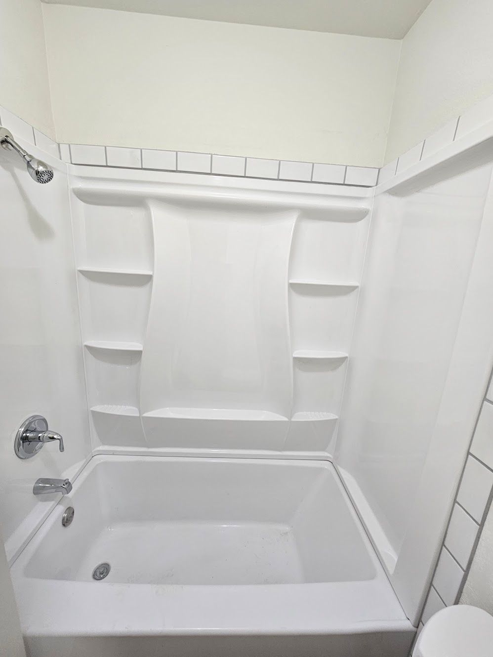 White bathroom, white tub with integrated shelves. Showerhead visible.