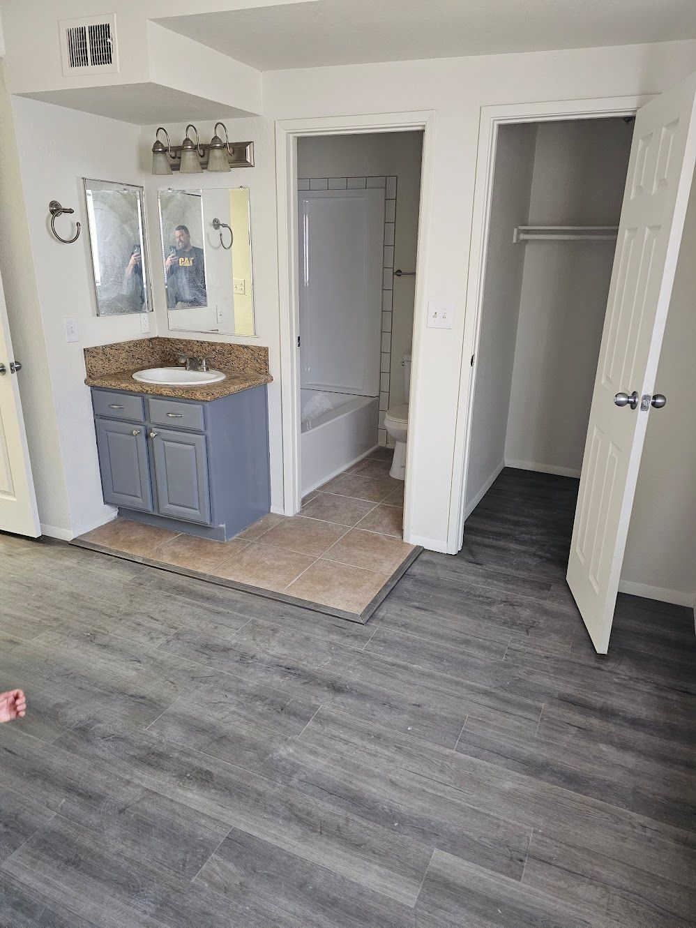 Bathroom and closet in room, gray cabinets, granite countertop, two mirrors, white door, gray flooring.