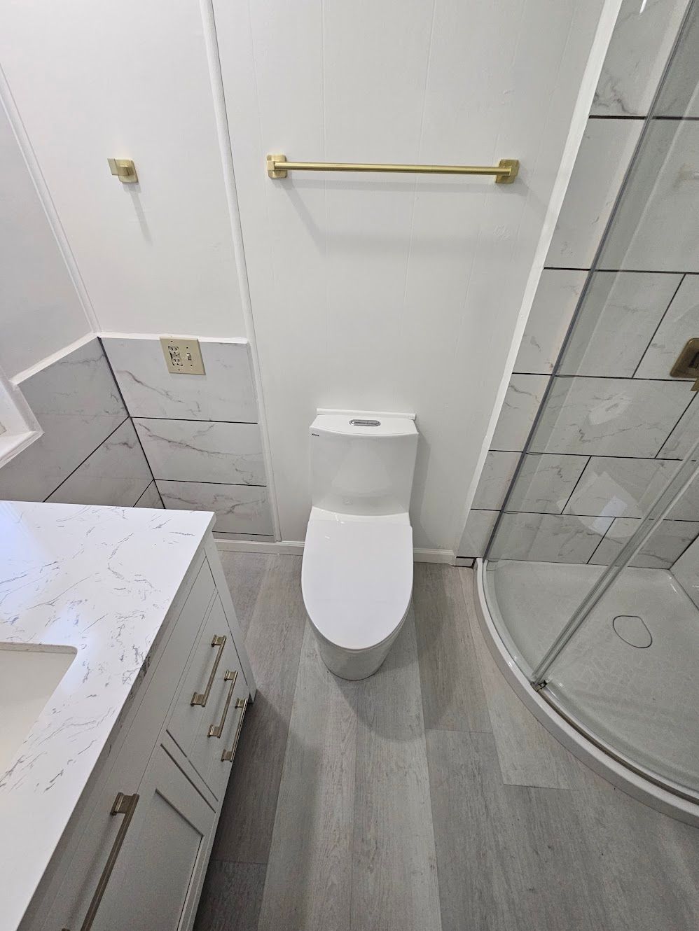 A modern bathroom with a white toilet, vanity, and shower. Gold towel bar and fixtures. Gray flooring.