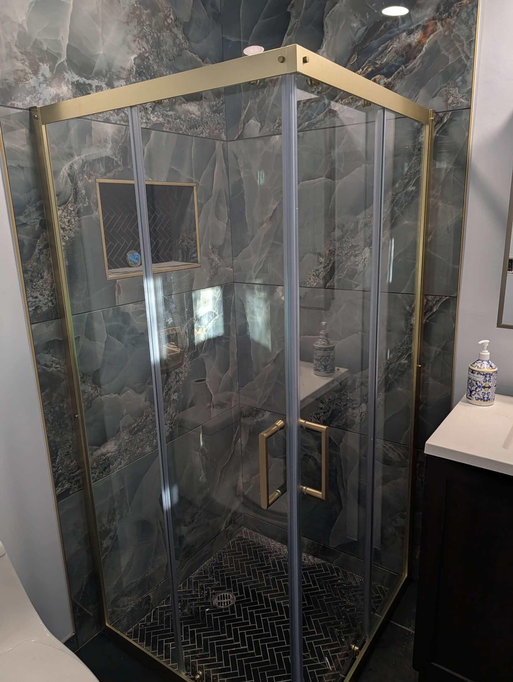 Gold-framed glass shower enclosure with blue-gray tile background and black pebble floor.