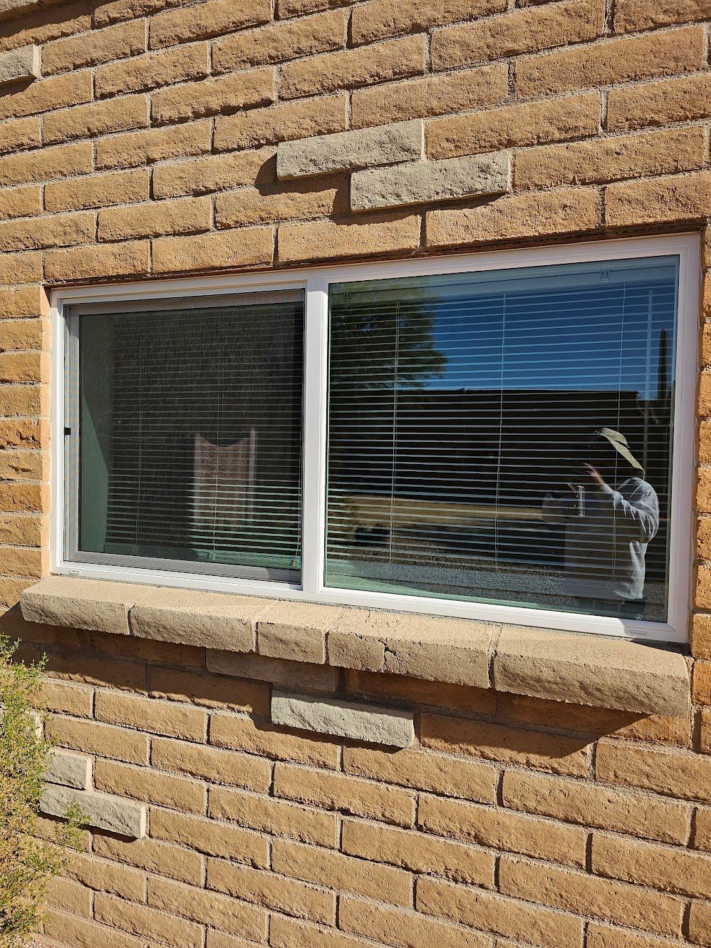White-framed sliding window with closed blinds in a brick wall.  Sunlight reflects in the glass.