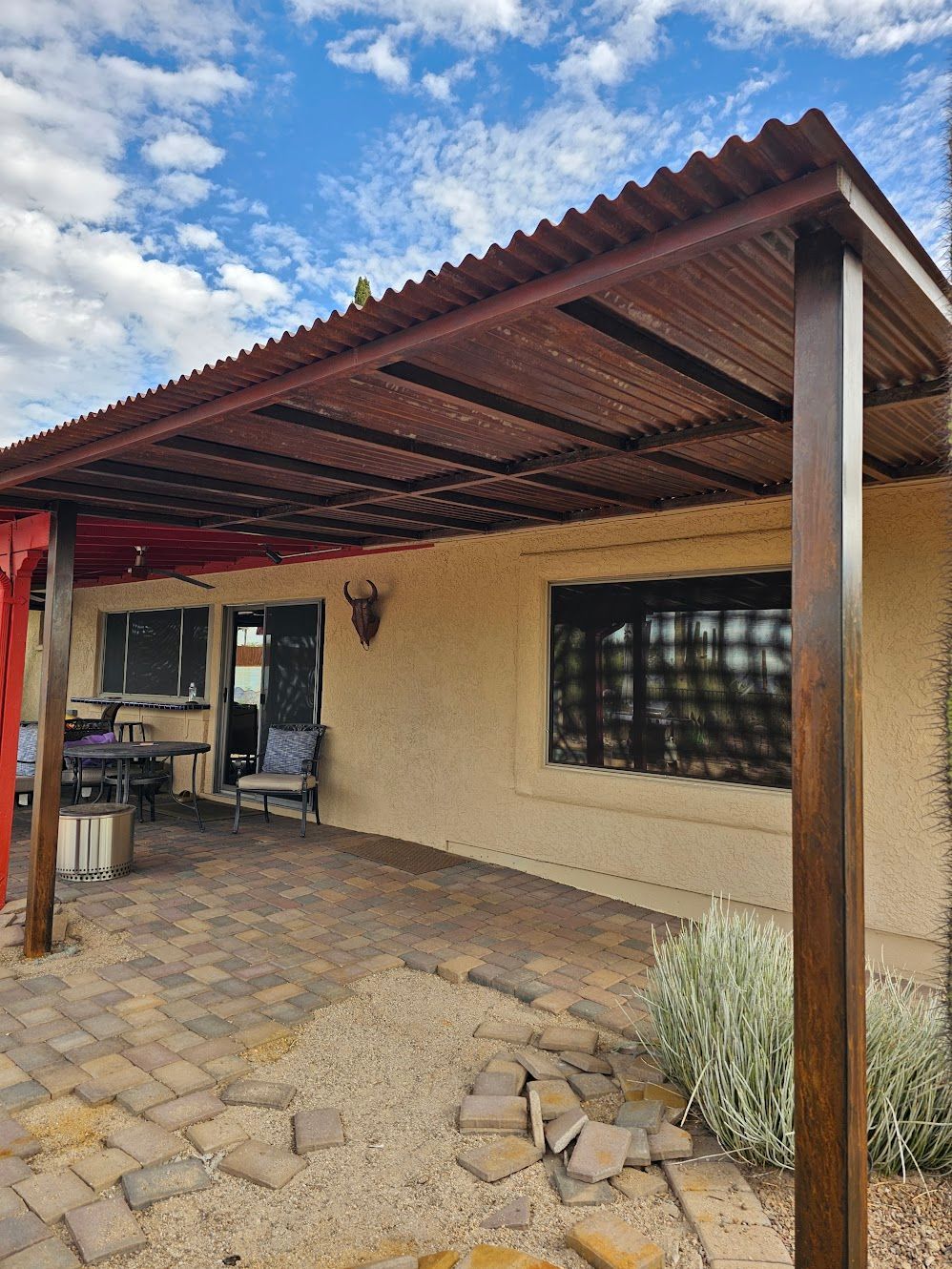 Patio with brown metal roof, brick patio, and stucco wall, under a blue sky.