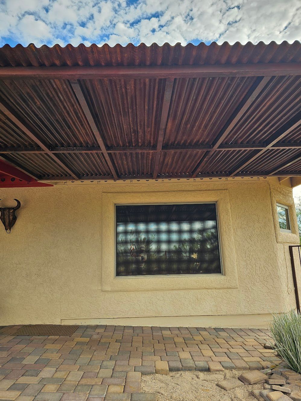 Tan stucco wall with a window, topped by a corrugated metal roof. A brick patio is in front.