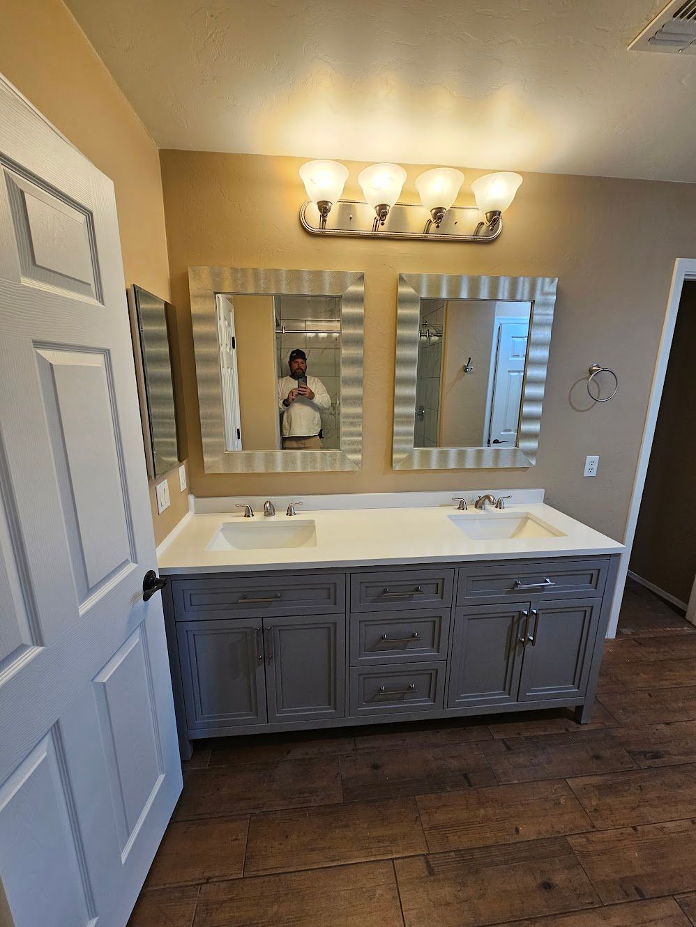 Bathroom with gray vanity, two mirrors, and overhead lighting. White sinks and door. Wooden floor.
