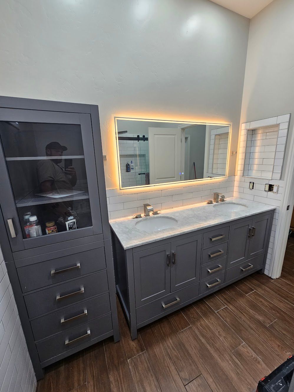 Gray bathroom vanity and cabinet with a lit mirror against a light-colored wall. Brown wood-look flooring.