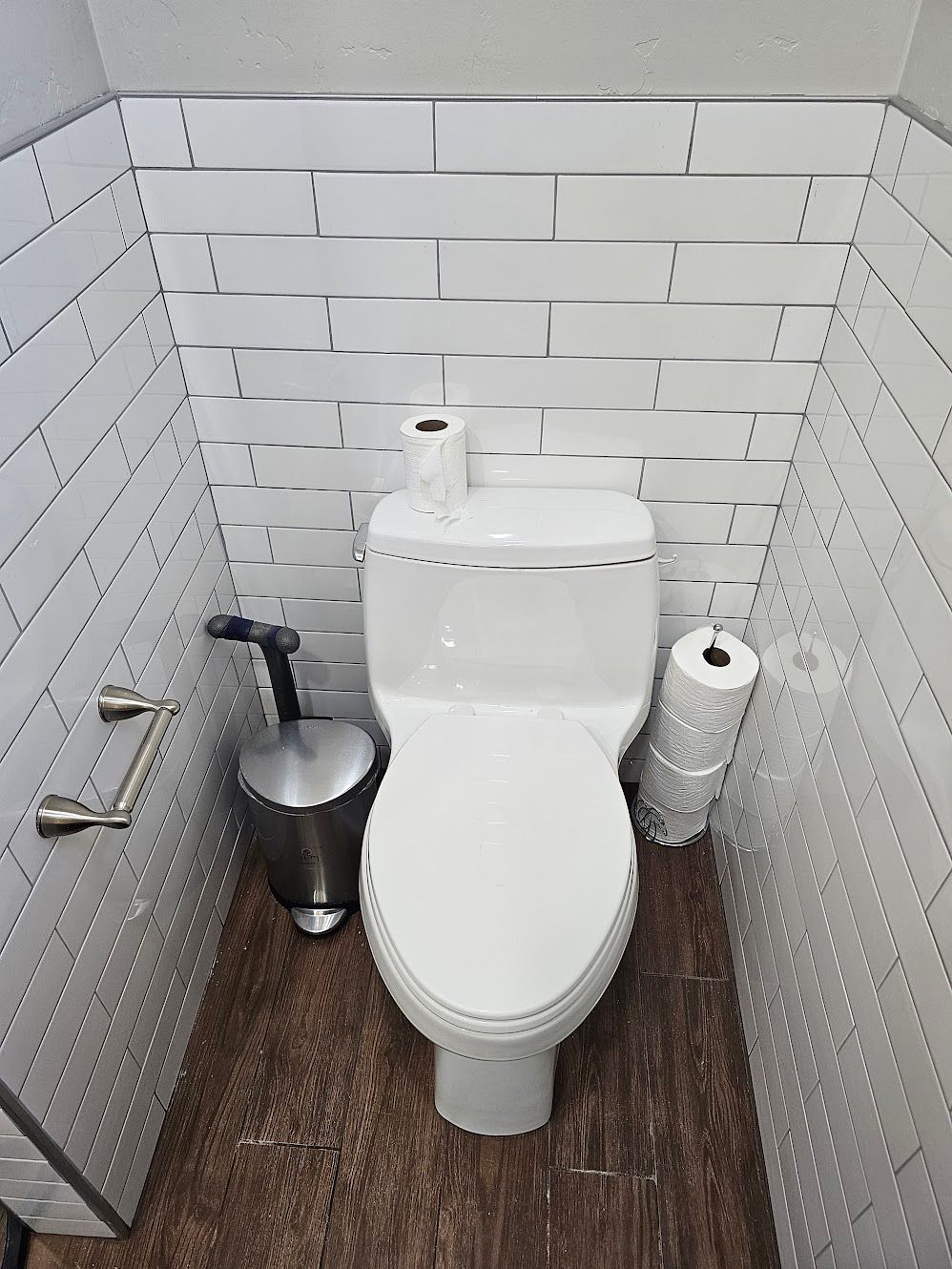 Toilet in a small, white-tiled bathroom nook with a wooden floor; a toilet brush and roll of toilet paper are nearby.