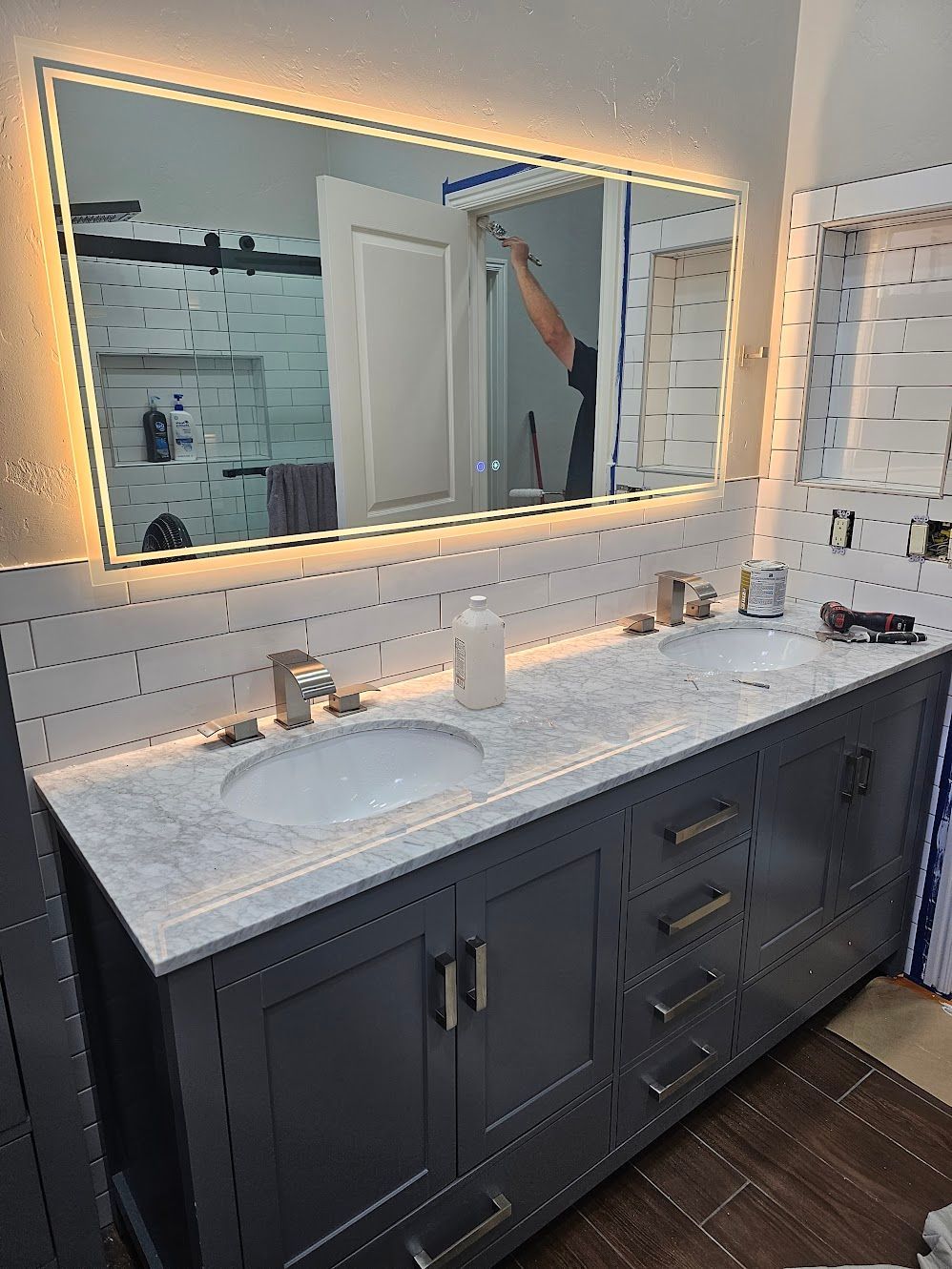 Gray double vanity with marble countertop, lit mirror, white tiled wall.