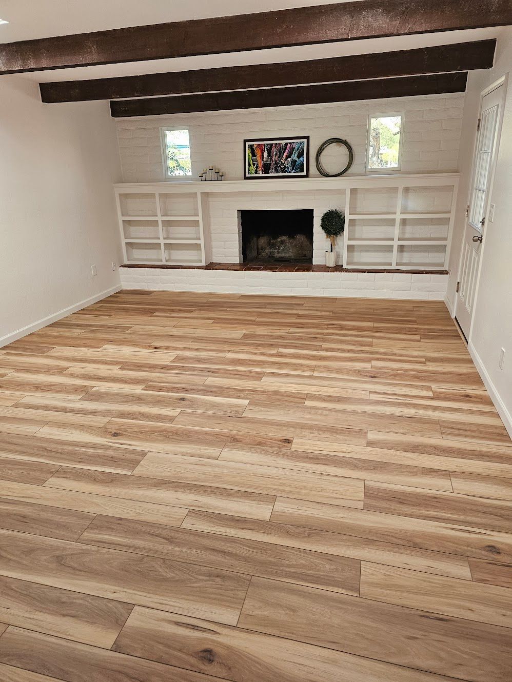Living room with light wood floors, white fireplace with built-in shelves, and dark wooden beams.