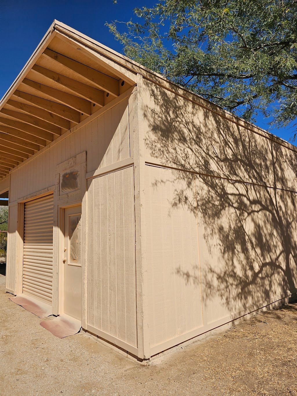 Tan-colored wooden building with a sloped roof. Sunlight casts shadows from nearby trees onto the side.