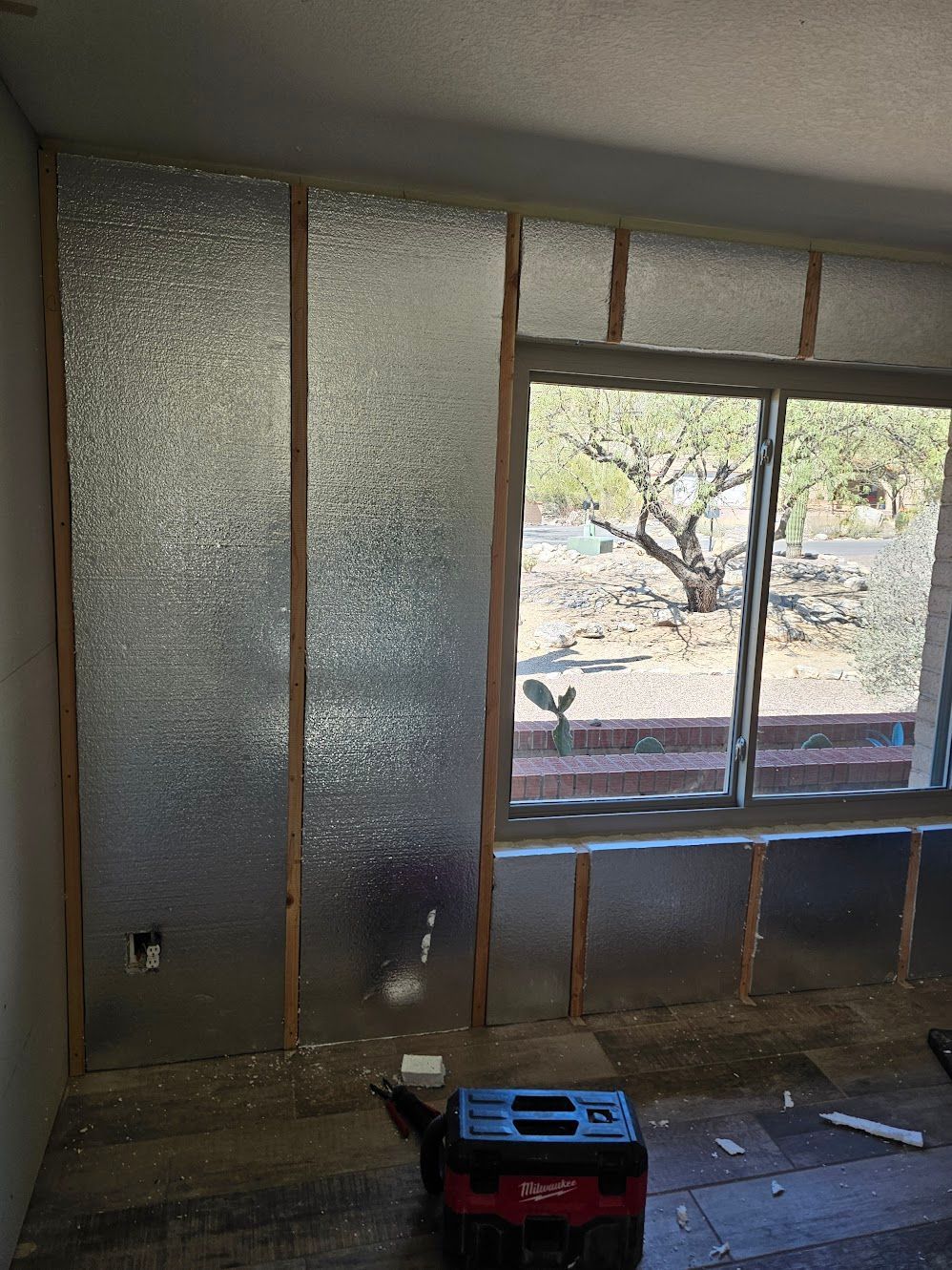 Interior wall under construction with foil insulation between wooden studs, next to a window with a tree view.