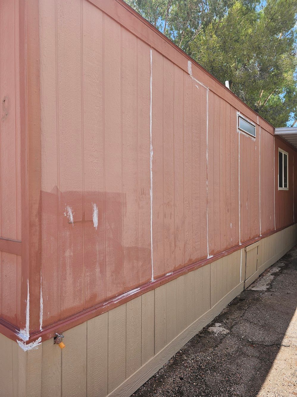 Side view of a reddish-brown mobile home with vertical paneling, light beige skirting, and a small window.