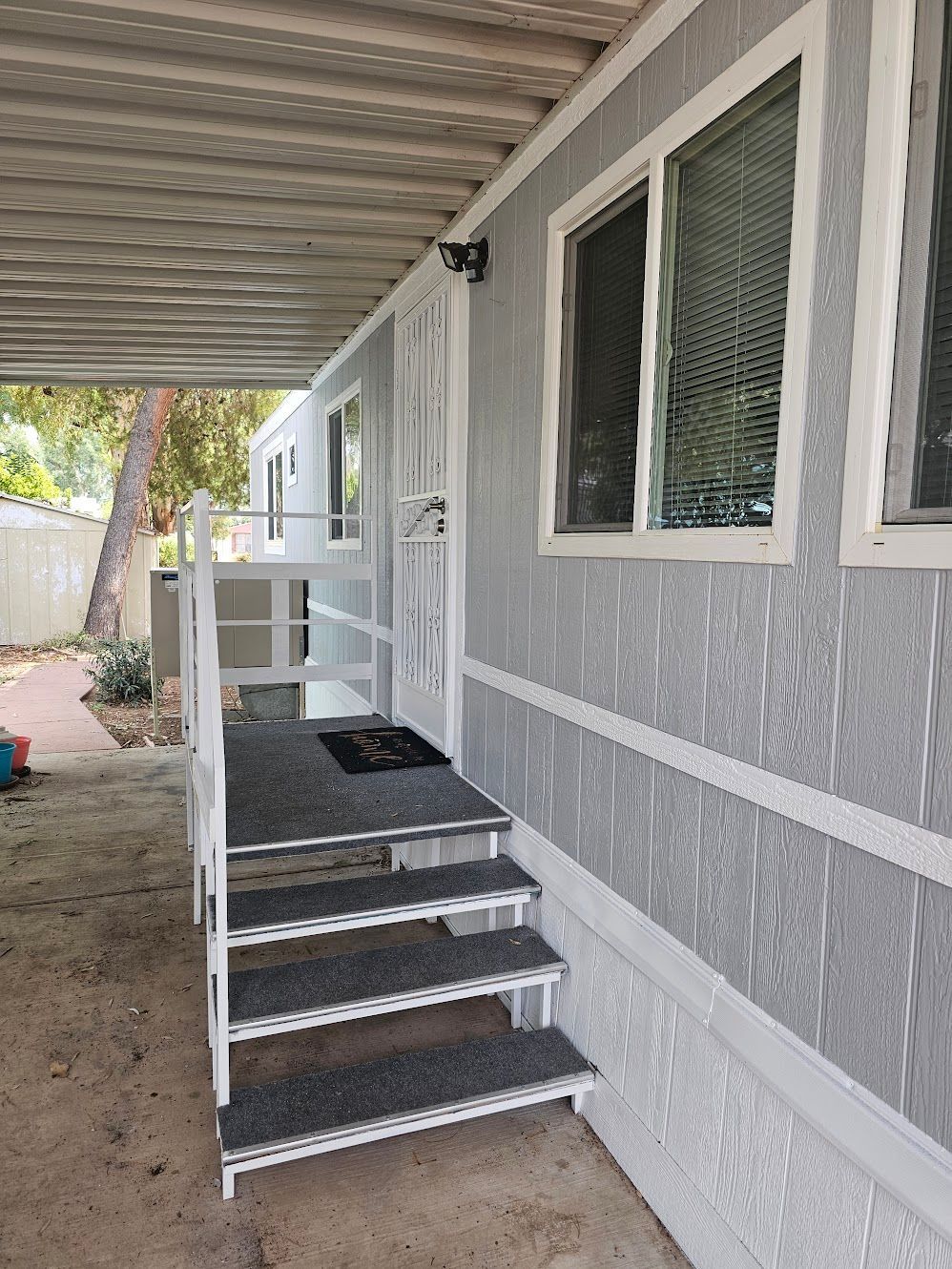 Exterior of a light grey mobile home. Steps lead to a screened door under a covered porch.