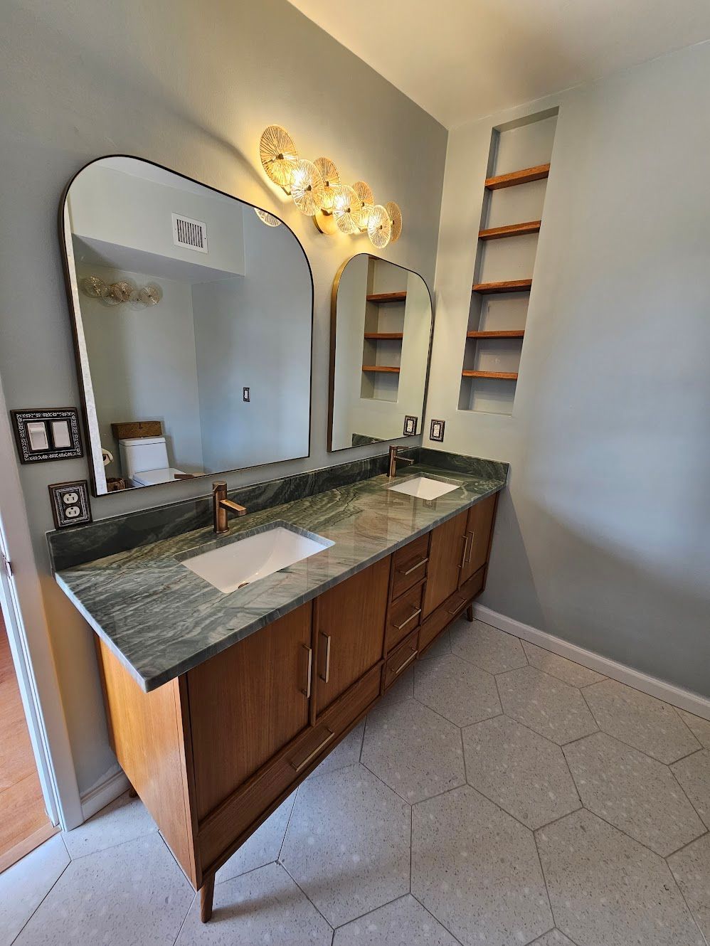 Bathroom with dual sinks, wooden vanity, green countertop, arched mirrors, and shelves.