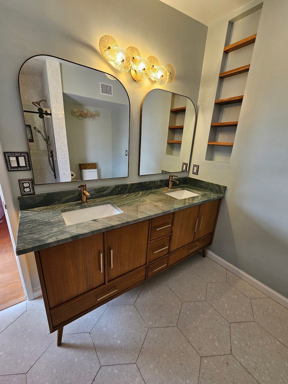 Bathroom with a wooden double vanity, two arched mirrors, gold light fixture, and green countertop.