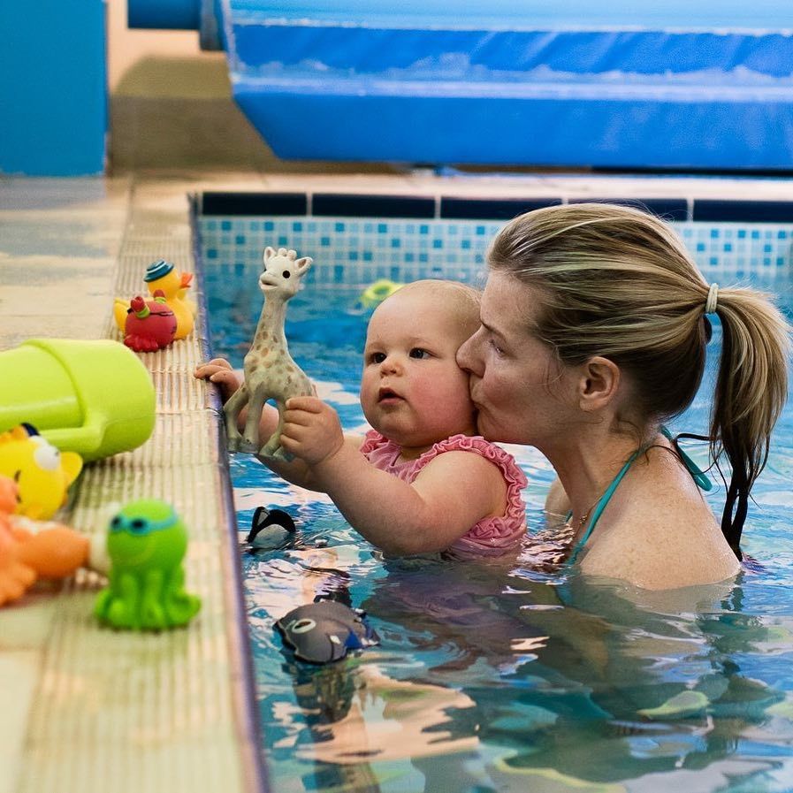 Woman Kisses Baby in Pool, Baby Holds Giraffe Toy, Surrounded by Colorful Bath Toys — Swim Care Boambee in Moonee, NSW