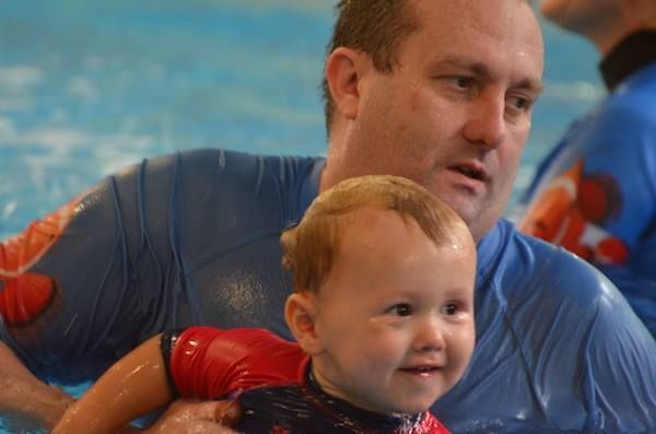 Man in Blue Shirt Holding Smiling Baby in Pool — Swim Care Boambee in Coffs Harbour, NSW