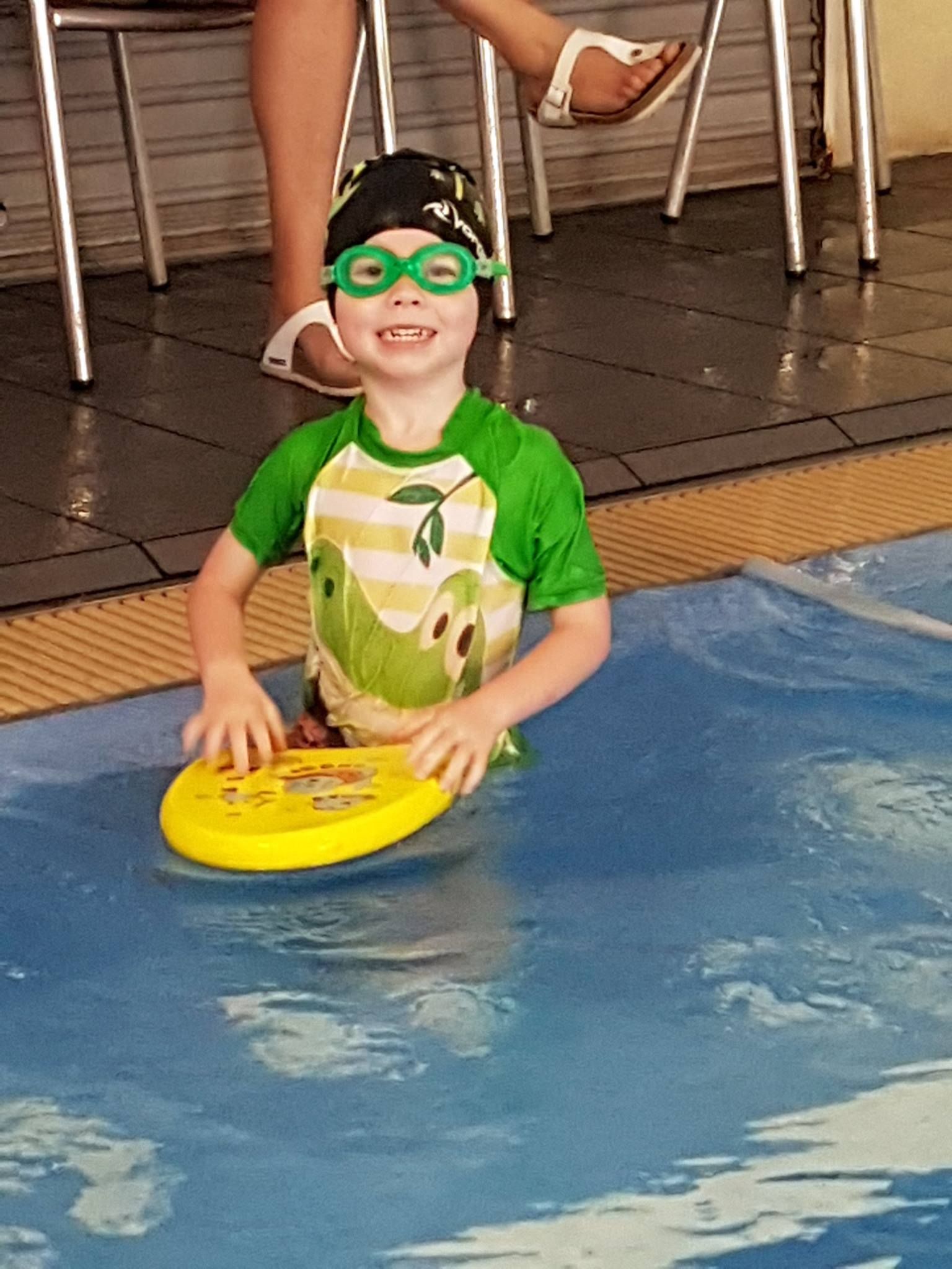 Boy in Green Swim Shirt and Goggles in Pool, Holding Yellow Kickboard, Smiling — Swim Care Boambee in Boambee, NSW