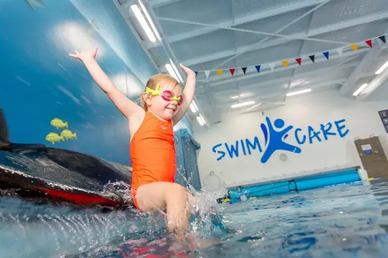Girl in Orange Swimsuit and Goggles Splashes in Indoor Pool With Swim Care Logo — Swim Care Boambee in Coffs Harbour, NSW