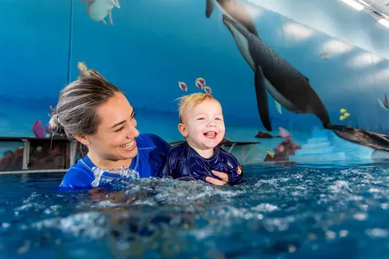 Woman and toddler in a pool, both smiling. The pool has an underwater mural.