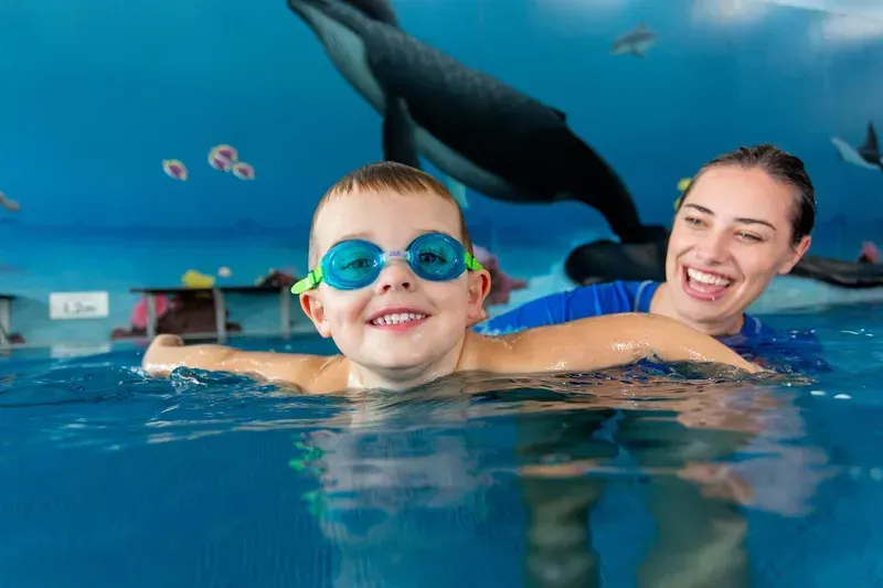 Boy With Goggles Smiles in Pool with Instructor, Whale Mural in Background — Swim Care Boambee in Coffs Harbour, NSW